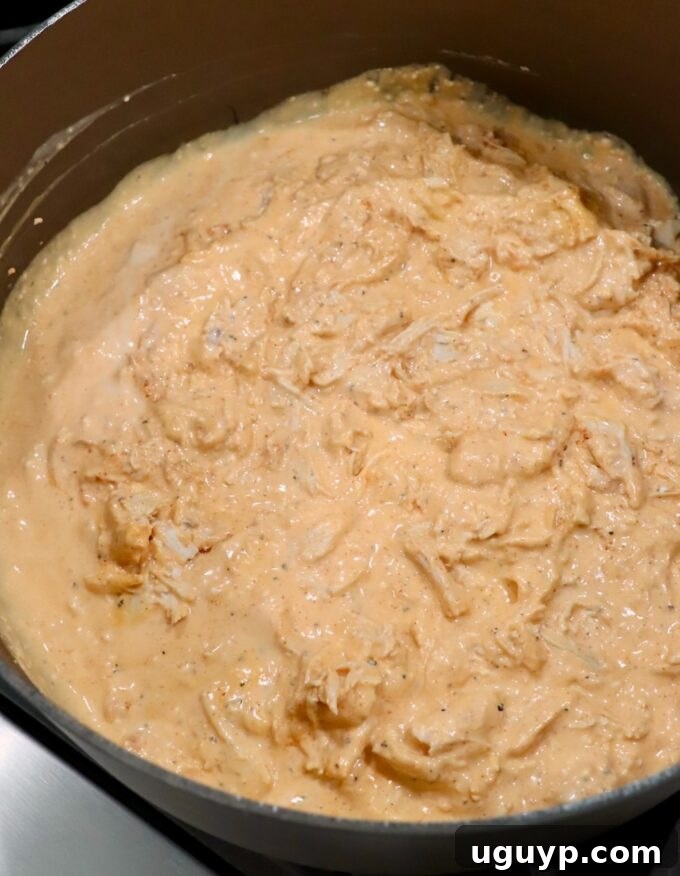 Cooked, shredded chicken being tossed into the creamy buffalo sauce in a large pot, integrating the flavors.