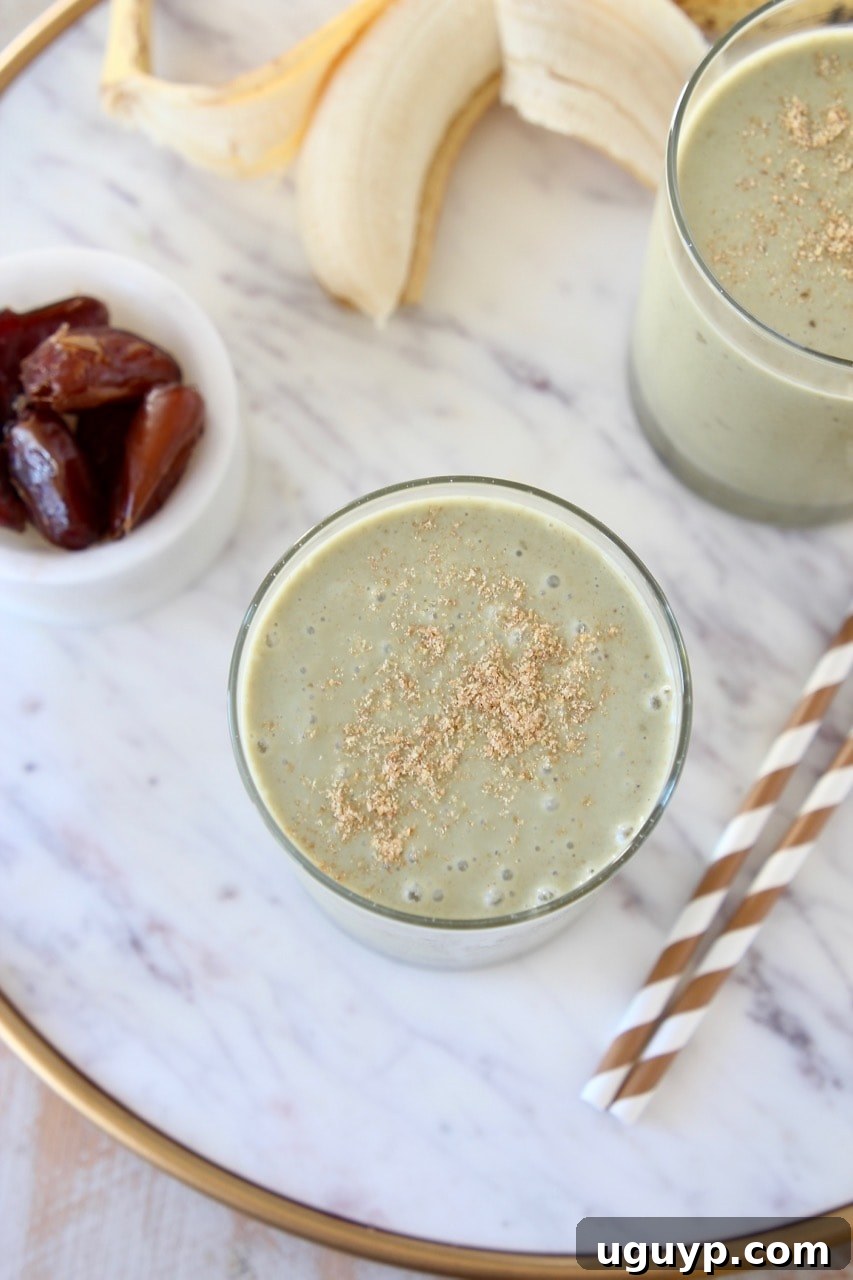 Overhead shot of two banana protein shakes topped with ground flaxseed on marble tray with gold and white striped straws and a small bowl of dates on the side
