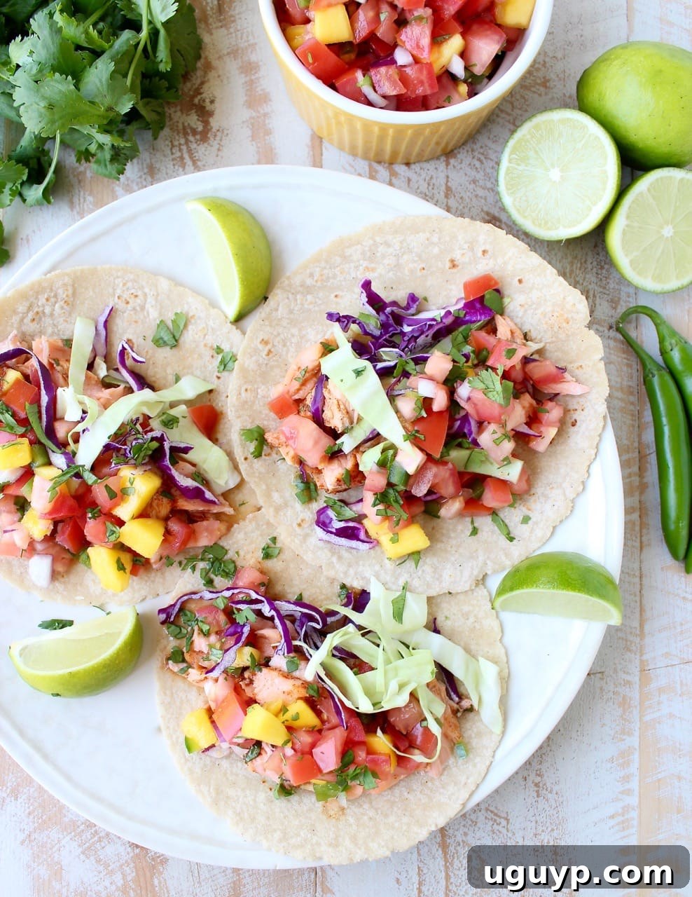 Three appetizing salmon tacos on a plate, garnished with fresh mango salsa and shredded cabbage, accompanied by lime wedges