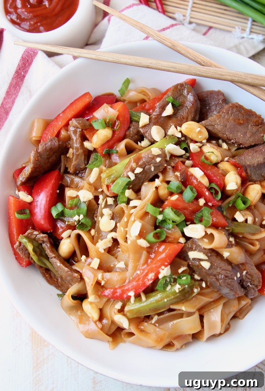 An overhead view of a bowl of Kung Pao Beef with rice noodles, garnished with green onions and bell peppers, chopsticks resting on the side.