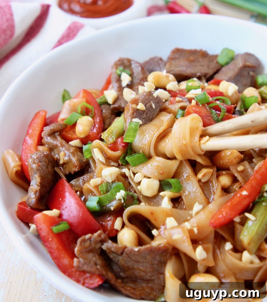 Close-up of chopsticks lifting a generous portion of Kung Pao noodles with beef, bell peppers, and peanuts from a bowl.