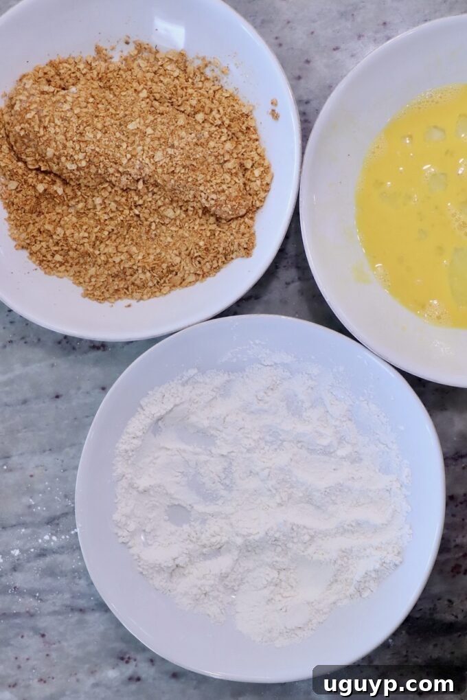 Three bowls on a marble surface contain ingredients for breading: flour, beaten eggs, and tortilla chip crumbs. The setup is ready for coating food.