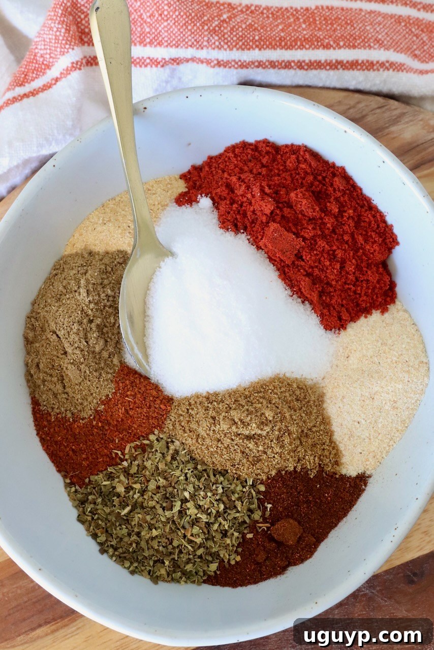A white bowl filled with colorful Al Pastor spices, including rich red chili powder, sparkling white salt, earthy brown cumin, and vibrant green oregano. A gold spoon rests elegantly on the side, inviting you to create culinary magic.
