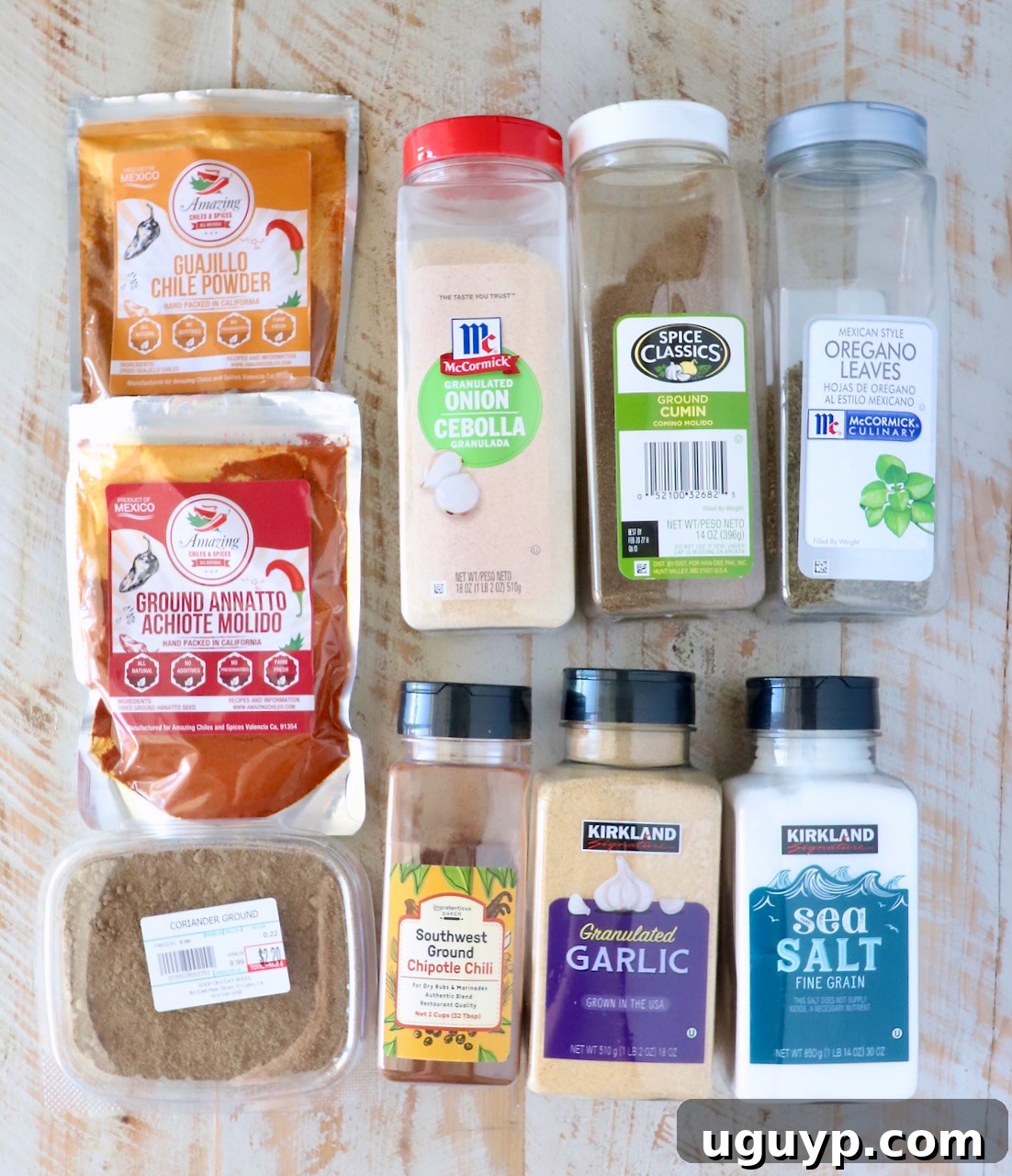 A variety of colorful seasoning packages are arranged neatly on a rustic wooden surface, featuring chili powders, granulated onion, cumin, oregano, garlic, sea salt, and more. This visual helps for your shopping list, showing the individual ingredients needed for a homemade Al Pastor seasoning blend.