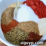 A white bowl filled with colorful Al Pastor spices, including rich red chili powder, sparkling white salt, earthy brown cumin, and vibrant green oregano. A gold spoon rests elegantly on the side.