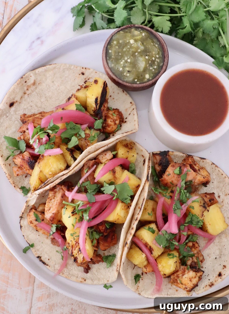 Three tacos filled with grilled chicken, pineapple, pickled onions, and cilantro on a plate. Beside them are bowls of green salsa and hot sauce.