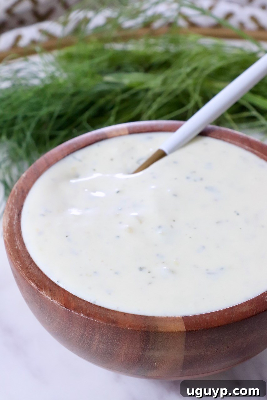 A wooden bowl filled with creamy, pickle ranch dressing, speckled with herbs. A white spoon rests inside. Fresh dill fronds are in the background.