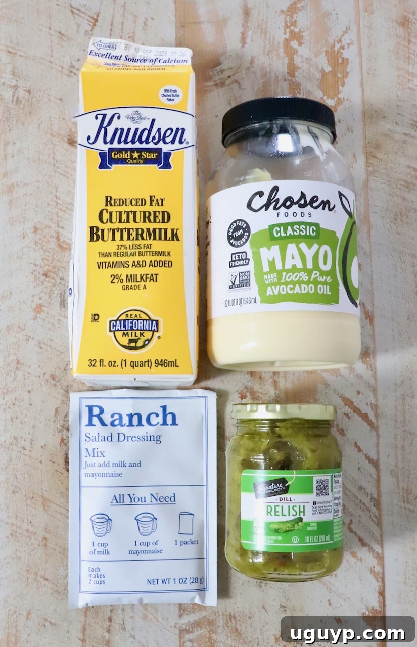 Four food items on a wooden surface: buttermilk carton, avocado oil mayo jar, ranch dressing mix packet, and dill relish jar.