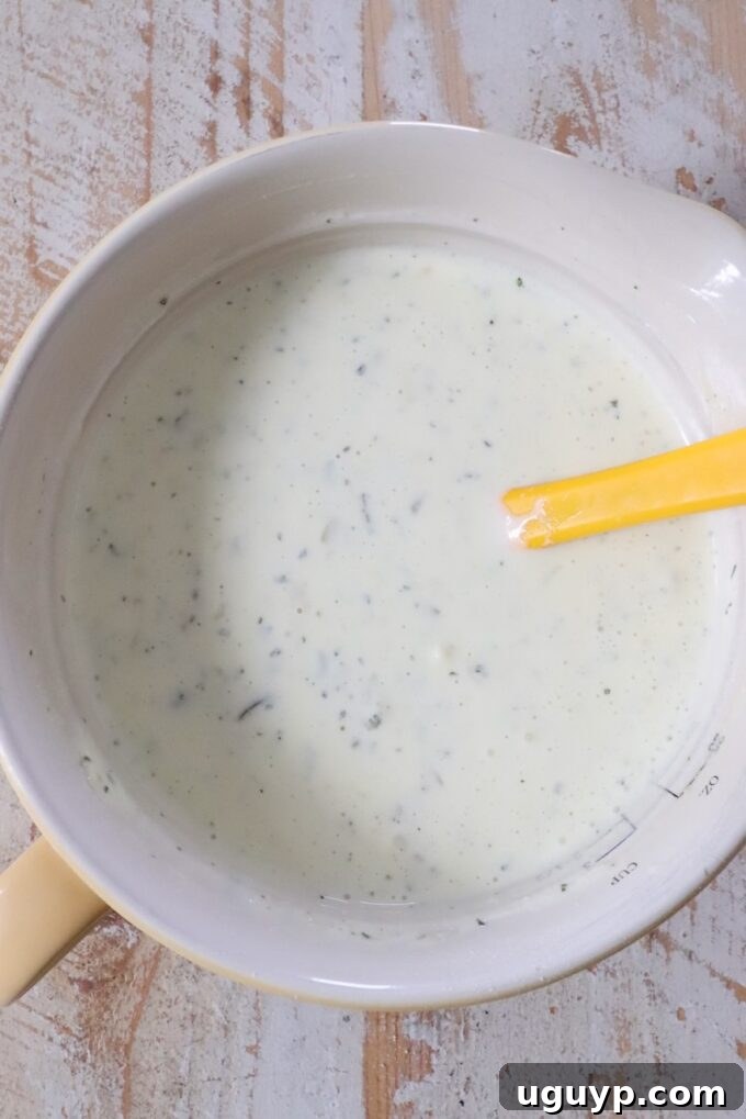 Creamy ranch dressing with herbs in a yellow mixing bowl, with a yellow spatula resting inside. 