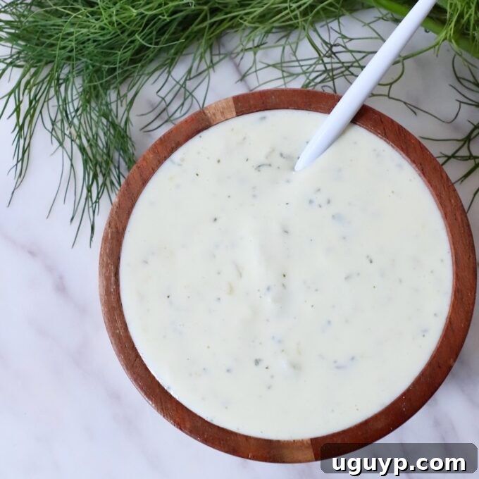 A wooden bowl filled with creamy, pickle ranch dressing, speckled with herbs. A white spoon rests inside. Fresh dill fronds are in the background.