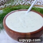 A wooden bowl filled with creamy, pickle ranch dressing, speckled with herbs. A white spoon rests inside. Fresh dill fronds are in the background.