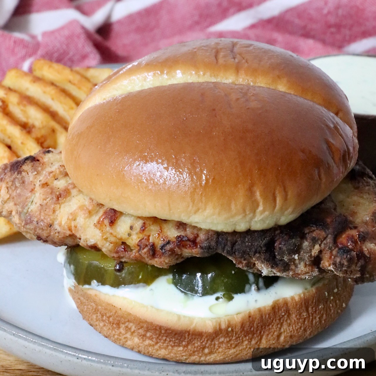 A crispy air fried chicken sandwich on a soft bun with pickles and creamy sauce, served with waffle fries on a plate.