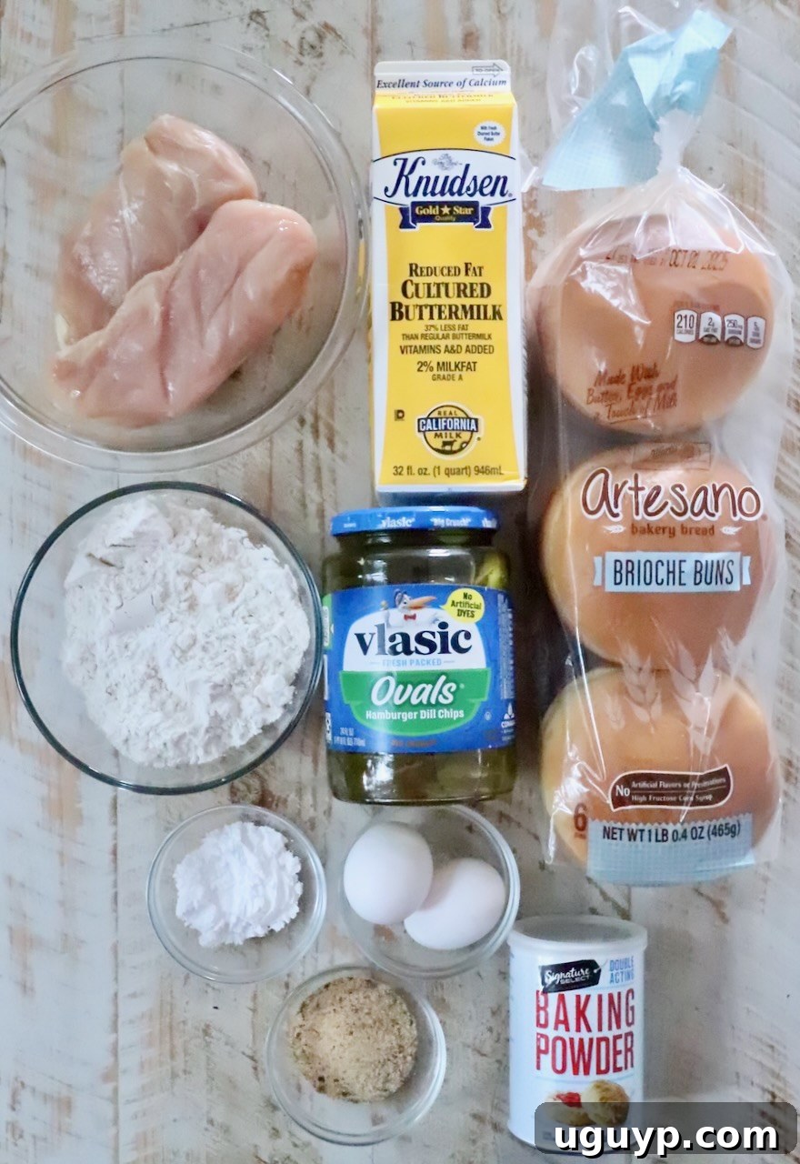 Ingredients for a chicken sandwich are neatly arranged: raw chicken, buttermilk, brioche buns, flour, pickles, eggs, baking powder, and chicken seasoning.