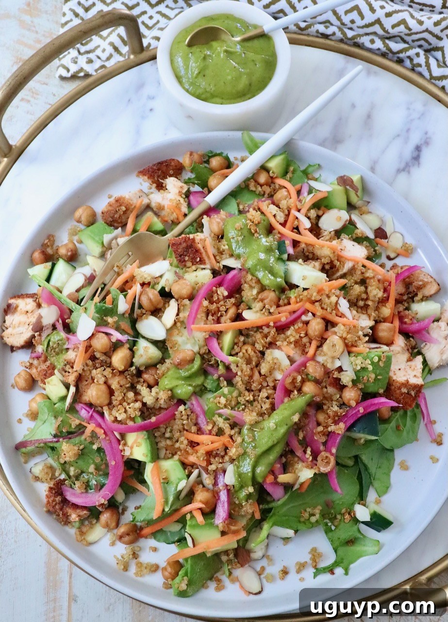A vibrant salad with greens, quinoa, chickpeas, sliced almonds, carrots, and pickled onions on a platter. A cup of green dressing is beside it, hinting at the delicious flavors within.
