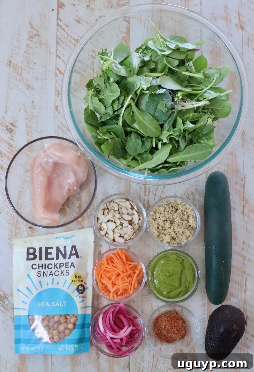 A top view of salad ingredients on a wooden surface, neatly arranged. Includes mixed greens, blackened chicken, diced cucumber, sliced avocado, crispy quinoa, sliced almonds, chickpea snacks, shredded carrots, and pickled red onions.