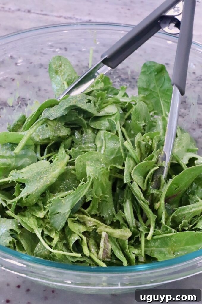 Mixed greens being tossed with vibrant green salad dressing in a large glass bowl with tongs, ensuring even coating.