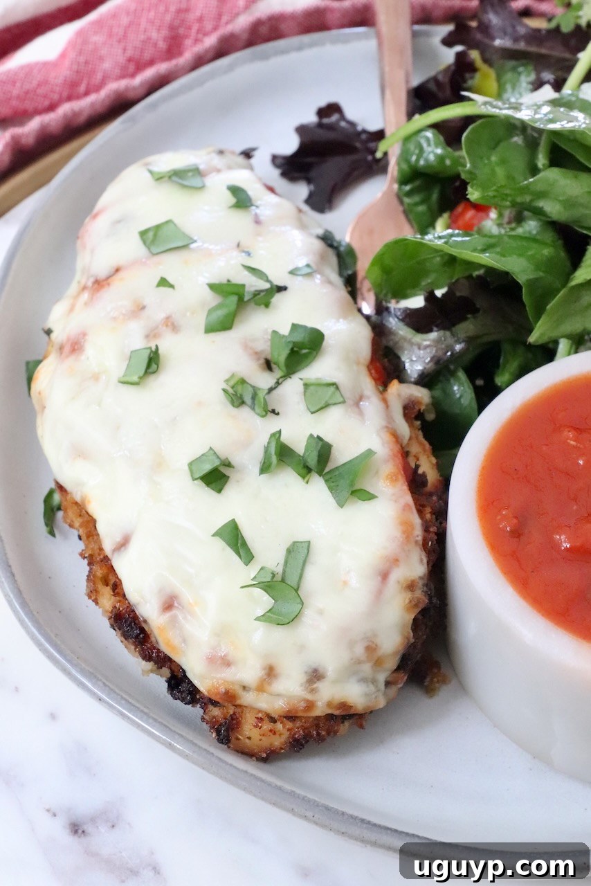 A delectable piece of Air Fryer Chicken Parmesan on a plate, served alongside a fresh green salad, with a fork and a small bowl of marinara sauce on the side.