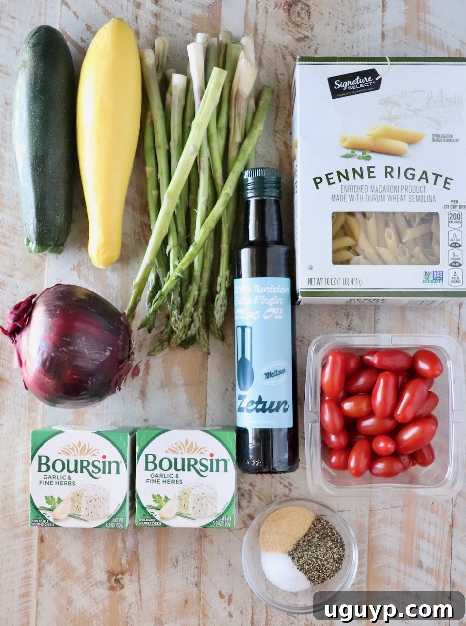 Assorted fresh ingredients on a wooden surface: zucchini, yellow squash, asparagus, red onion, cherry tomatoes, penne pasta, olive oil, Boursin cheese, and spices.