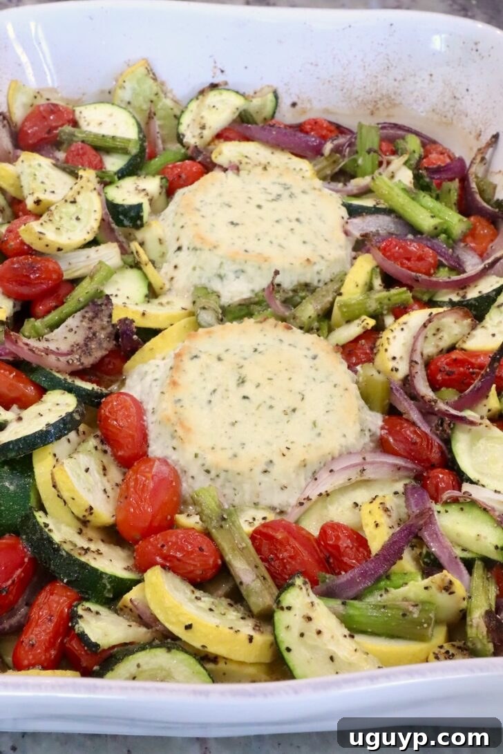 Roasted vegetables with two rounds of baked boursin cheese in a white baking dish. The veggies include zucchini, cherry tomatoes, asparagus, and red onions, seasoned with salt and pepper.