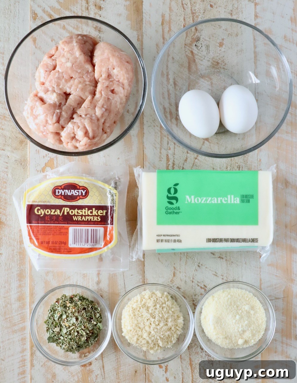 A top-down view of cooking ingredients on a wooden surface: ground chicken, two eggs, gyoza wrappers, mozzarella cheese, herbs, breadcrumbs, and grated cheese in bowls.