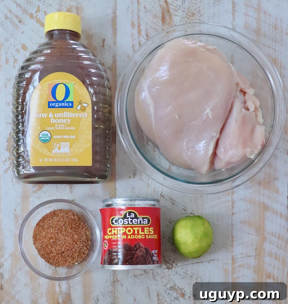Ingredients for cooking are arranged on a wooden surface: raw honey in a bottle, raw chicken breasts in a bowl, chipotle peppers in a can, a lime, and a bowl of taco seasoning.