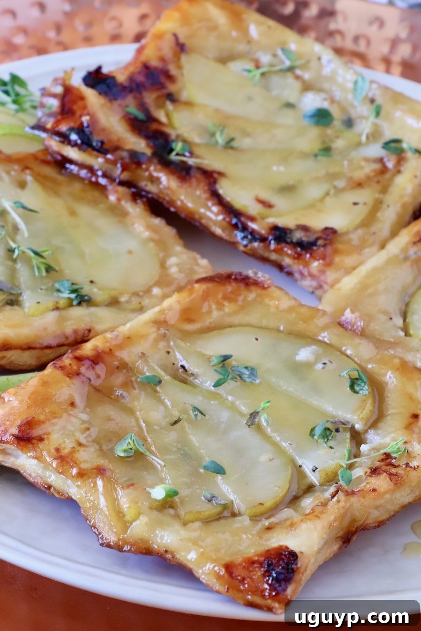 Close-up of golden-brown puff pastry tarts topped with sliced pears and drizzled with honey, garnished with fresh thyme leaves.