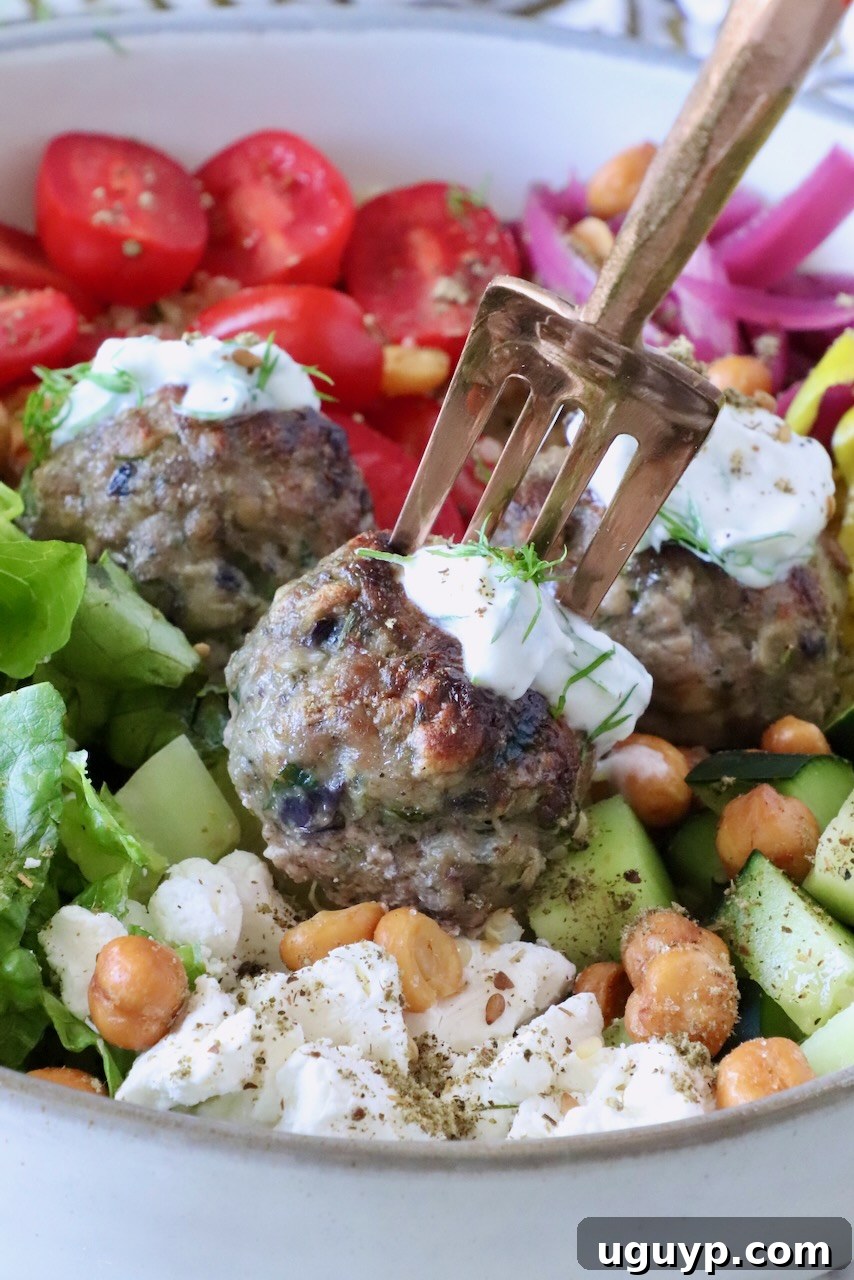 A bowl of vibrant Mediterranean salad with gyro meatballs topped with tzatziki sauce, sliced cherry tomatoes, cucumbers, crispy chickpeas, feta, and greens.