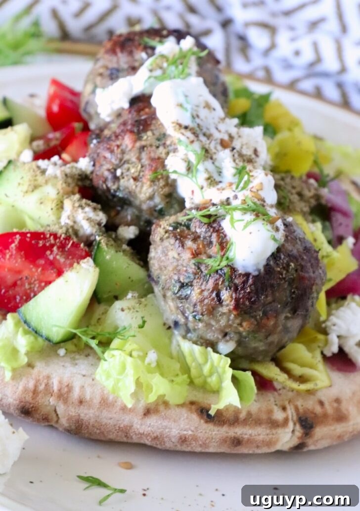 meatballs on top of pita bread on a plate, filled with lettuce, tomatoes, cucumber, and topped with creamy tzatziki sauce