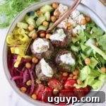 A colorful Mediterranean bowl with meatballs topped with yogurt sauce, surrounded by fresh lettuce, cherry tomatoes, cucumber, onions, and chickpeas.
