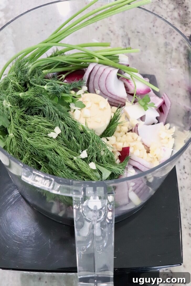 A food processor bowl filled with fresh herbs, sliced red onions, garlic, and parsley on a kitchen counter.