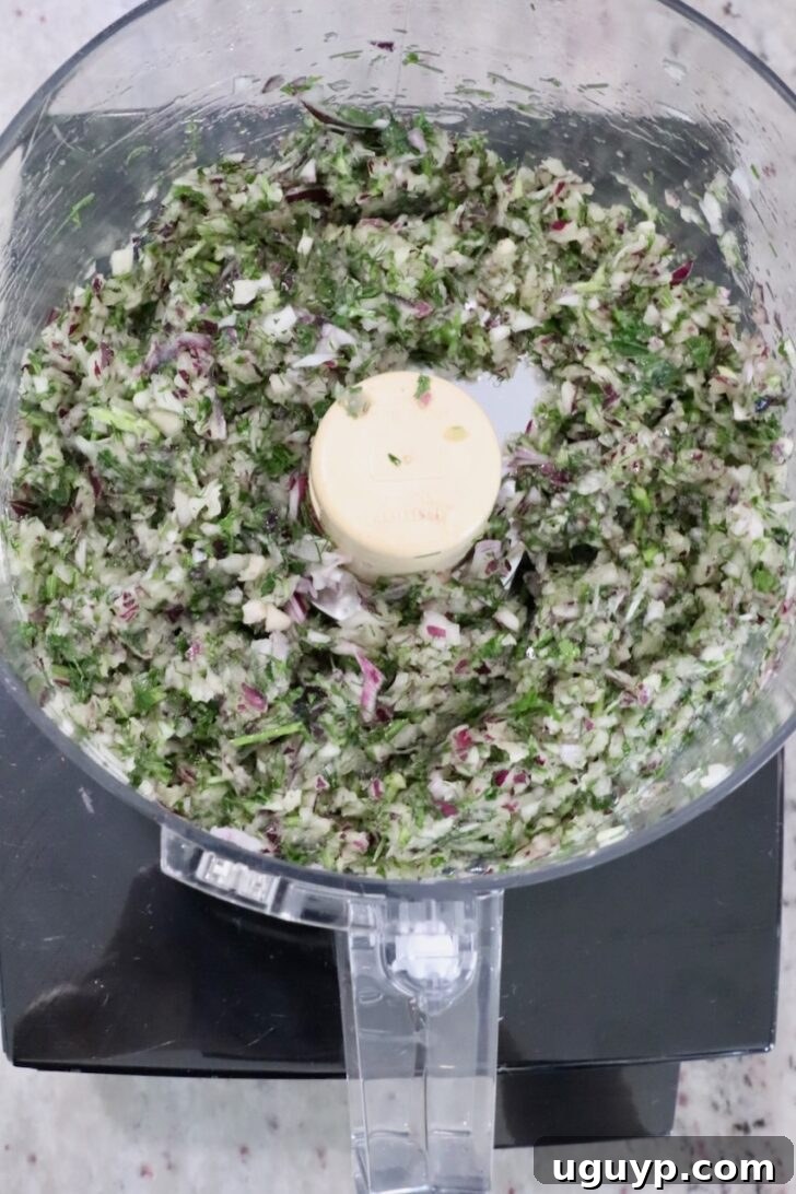 Chopped herbs and vegetables, including red onion and parsley, in a food processor bowl.
