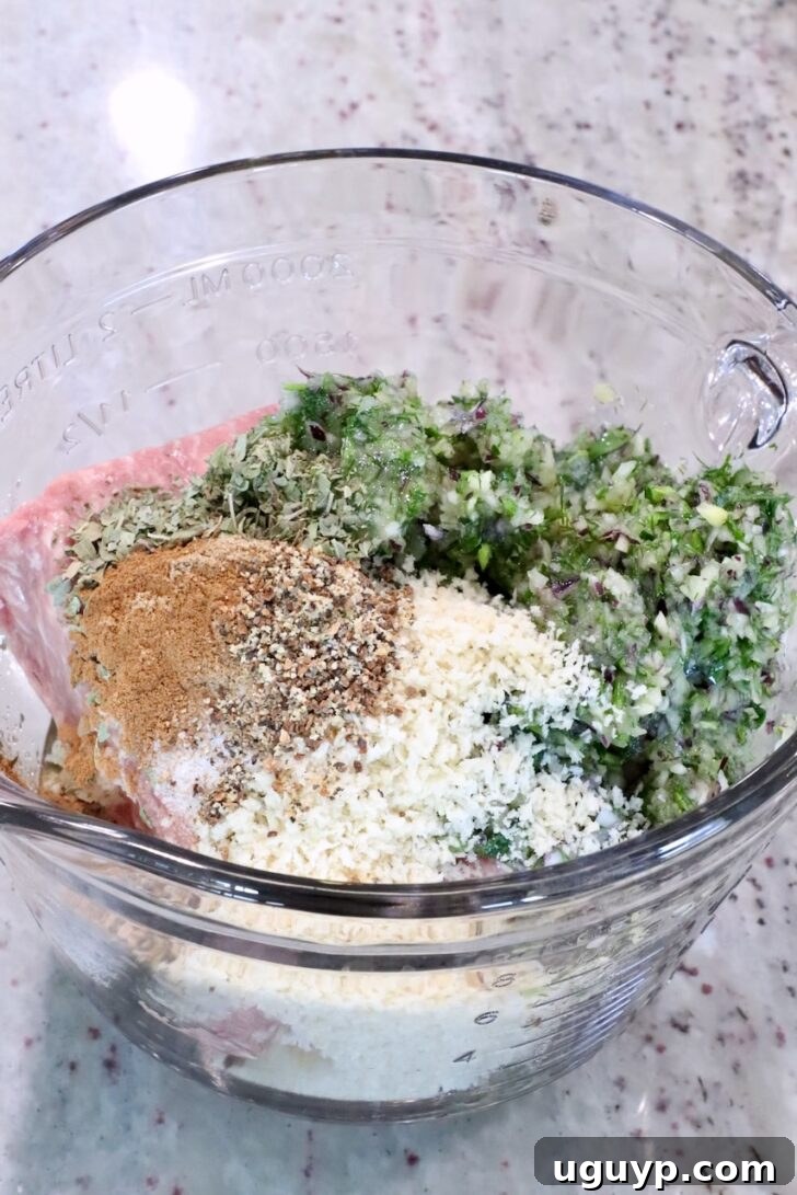 Glass bowl with minced meat, breadcrumbs, chopped herbs, and spices on a marble countertop.