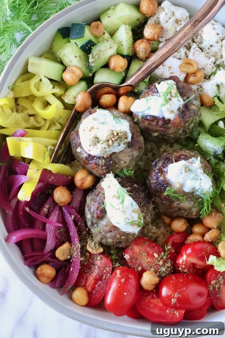 bowl filled with gyro meatballs topped with tzatziki sauce, surrounded by cucumbers, tomatoes, pickled onions, and chickpeas