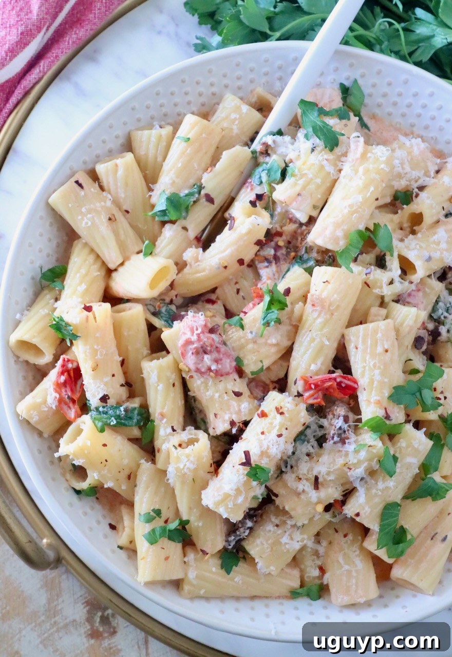 A bowl of creamy rigatoni pasta topped with fresh parsley, grated cheese, and red pepper flakes, mixed with sun-dried tomatoes; a fork rests in the bowl.
