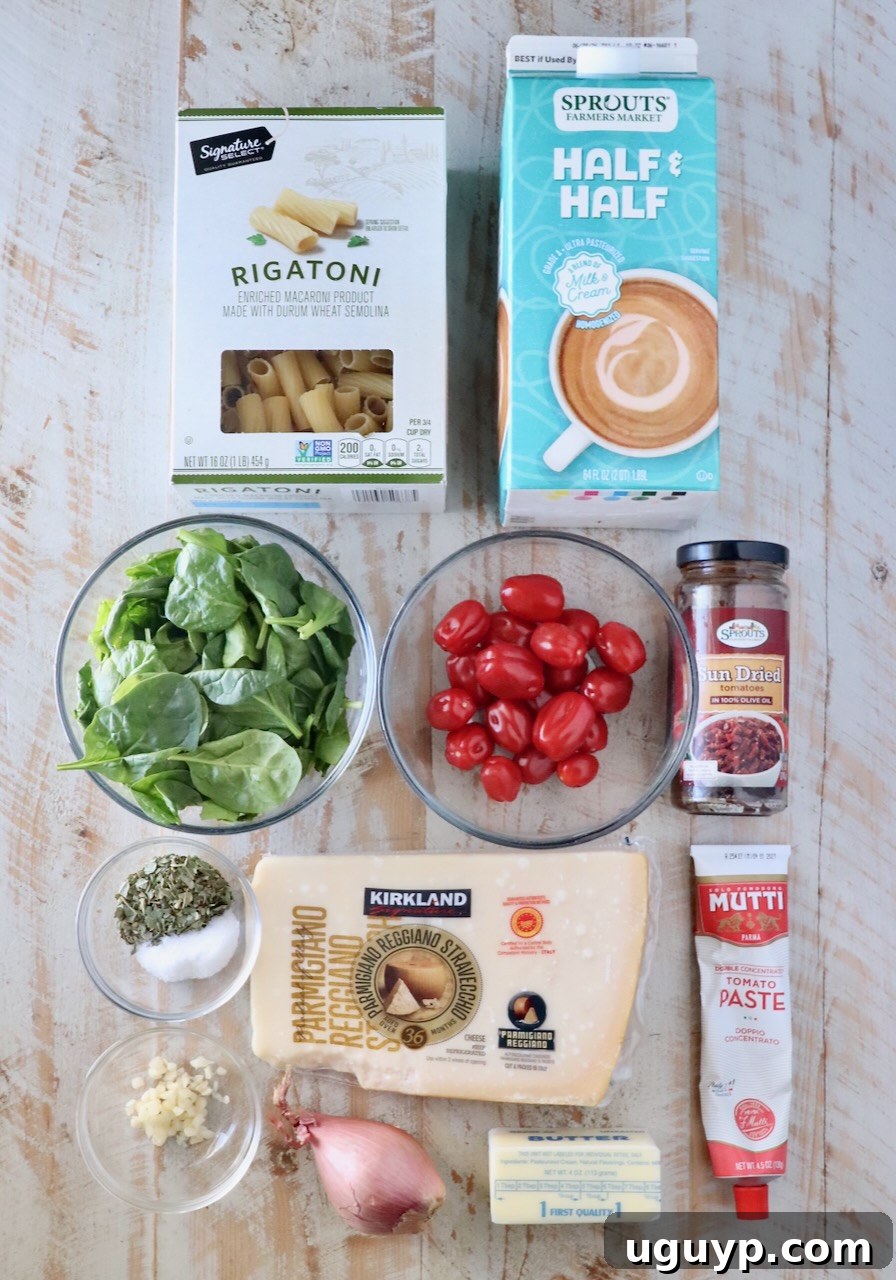 Assorted ingredients on a light wooden surface, including rigatoni, half & half, spinach, cherry tomatoes, garlic, herbs, parmesan, tomato paste, shallot, butter, and sun-dried tomatoes.
