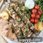 grilled chicken souvlaki on a plate with pita bread, a small bowl of tzatziki sauce, tomatoes, lemons, and herbs