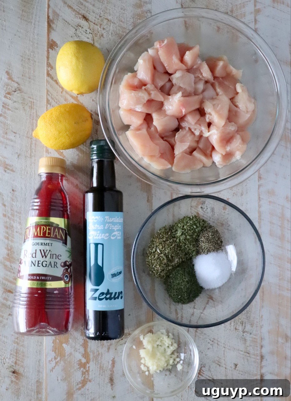 ingredients for souvlaki on a white wood board including a bowl of cubed chicken, olive oil, lemons, red wine vinegar, minced garlic, and spices