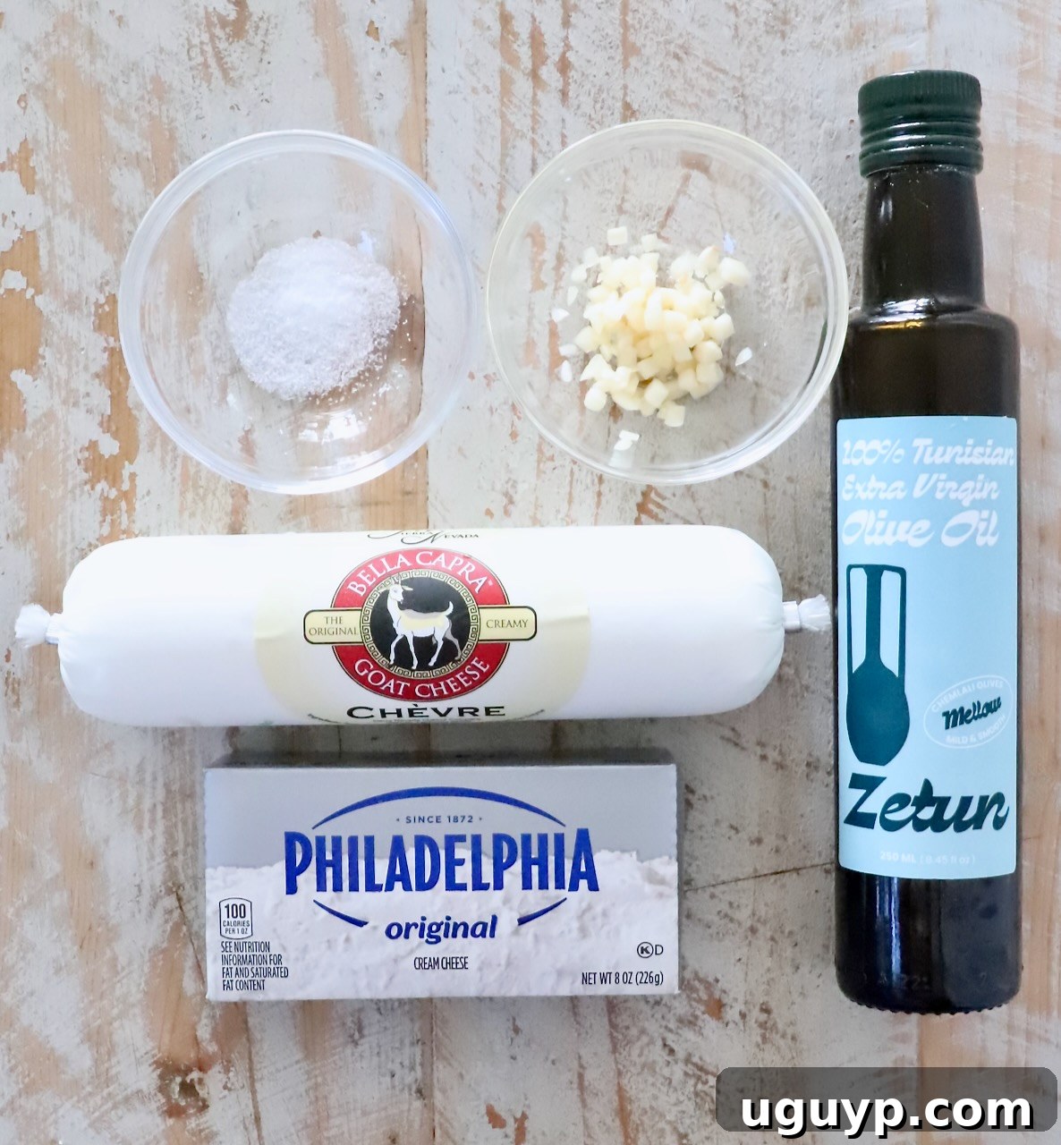 ingredients for goat cheese dip on a white wood board including olive oil, cream cheese, goat cheese, minced garlic and salt