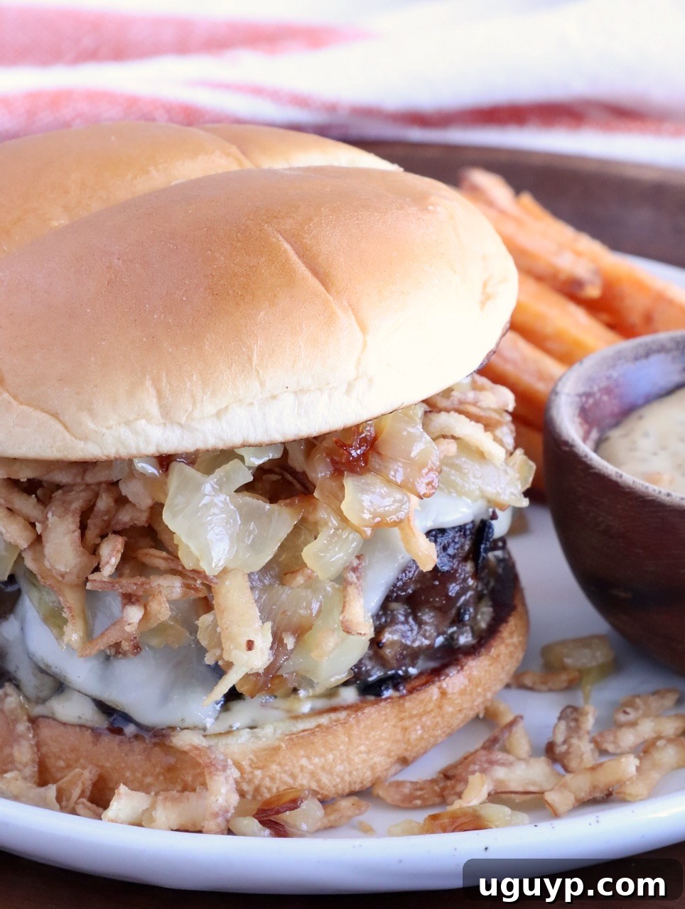 cheeseburger on a plate topped with crispy onions, with a side bowl of sauce, and sweet potato fries behind the burger on the plate