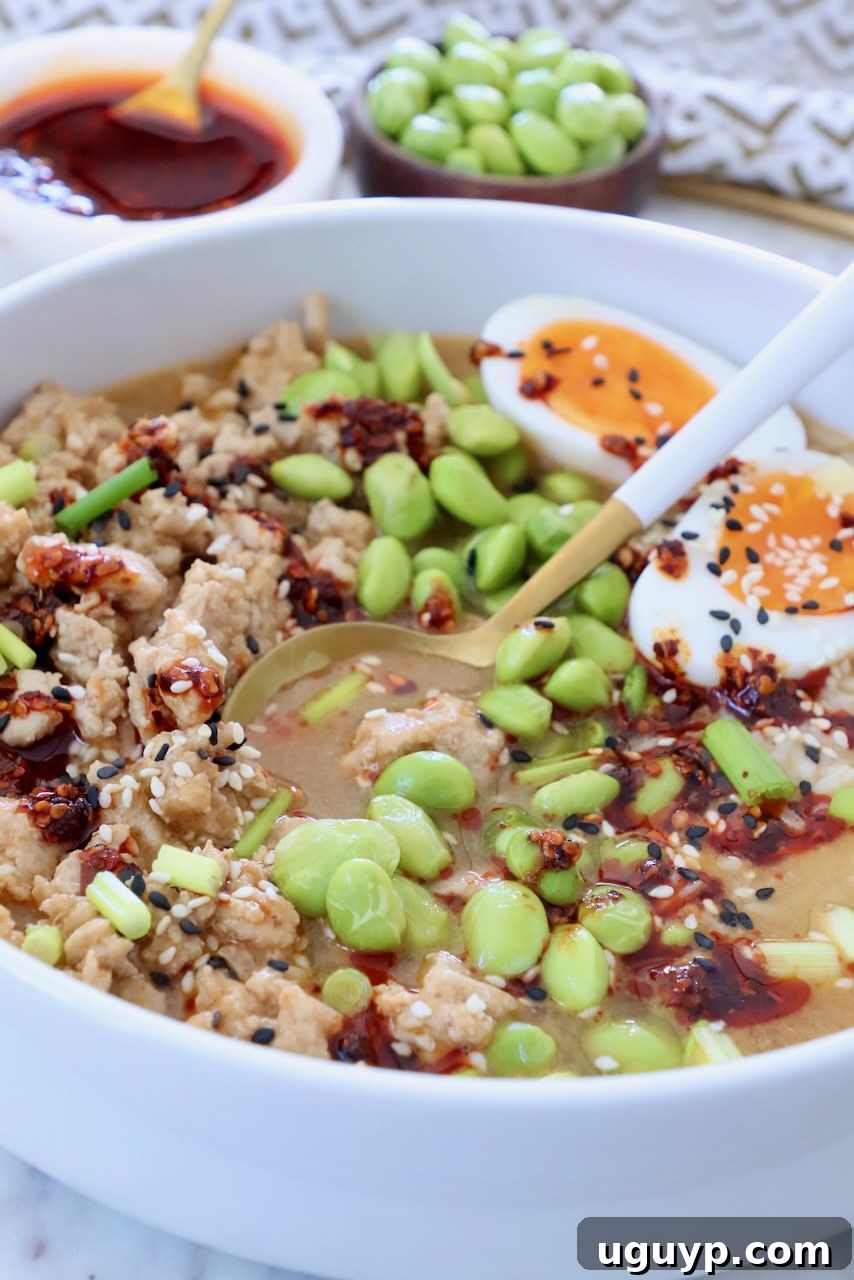 chicken broth bowl with edamame, chili crisp, and a soft-boiled egg cut in half, garnished with sesame seeds and green onions