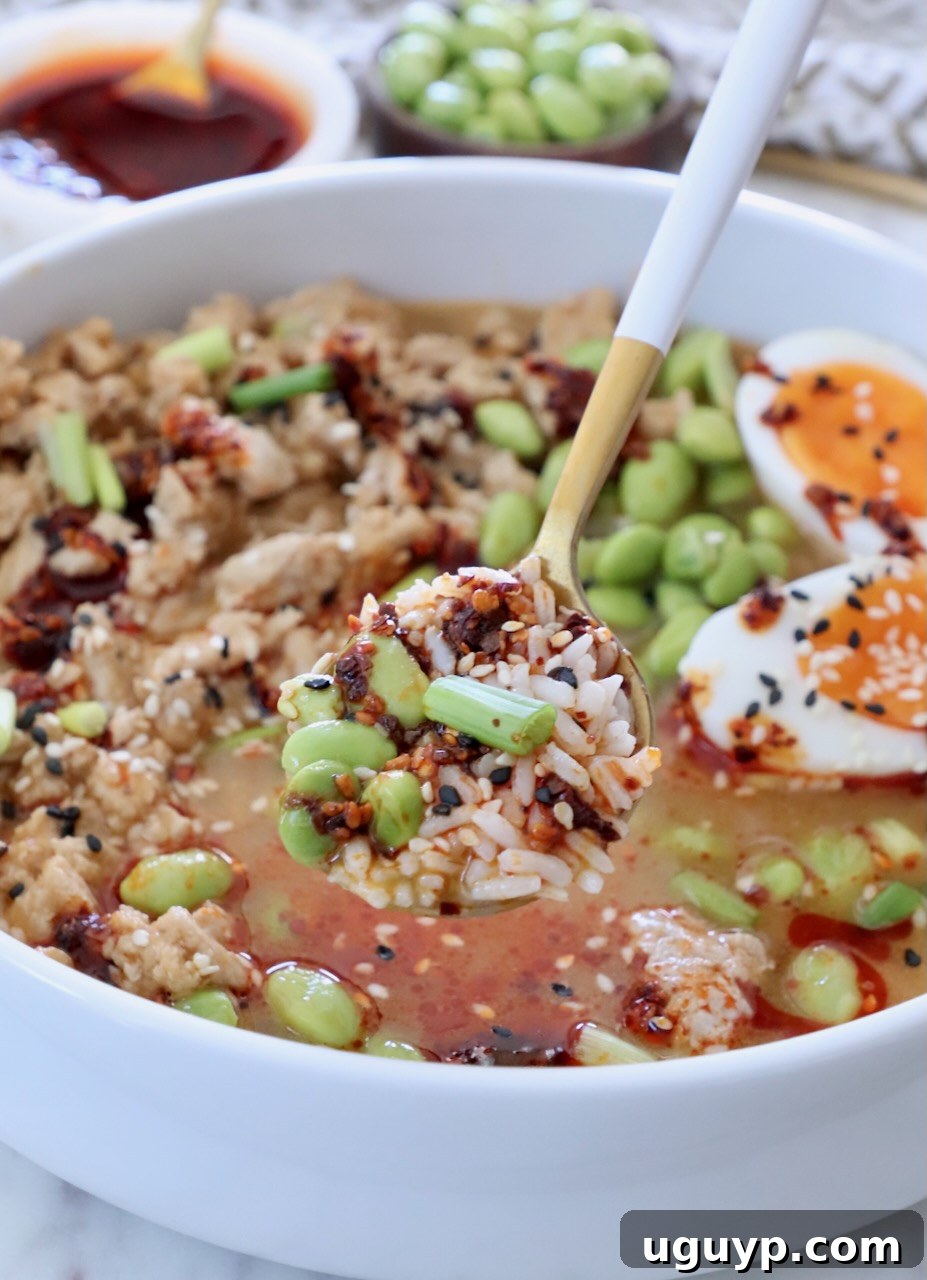 spoon scooping rice and edamame from a broth bowl with ground chicken, a jammy egg, and chili crisp on top